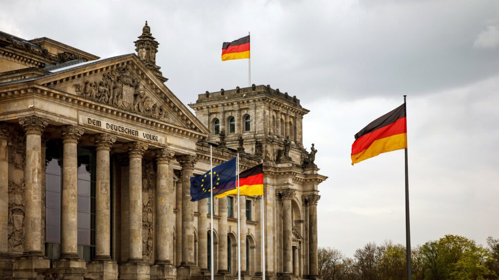Photo of the Bundestag building, with German flags flying beneath a cloudy sky