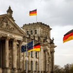 Photo of the Bundestag building, with German flags flying beneath a cloudy sky