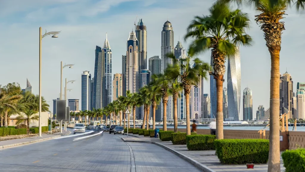 Seaside promenade on Palm Jumeirah, with palm-lined roads and light traffic before sunset, and the Dubai Marina towers visible in the background, in Dubai, United Arab Emirates.