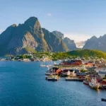 Scenic view of Reine village in the Lofoten Islands, showing traditional red and white houses along the shoreline surrounded by steep mountains and clear blue water.