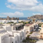 Muscat, Oman, with white buildings spread across the capital city and surrounded by rugged mountains and dramatic rocky landscapes.